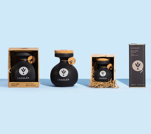 Three LADOLEA products arranged on a light blue studio background: a boxed olive oil set, an unboxed dark bottle with cork stopper and gold tag, a boxed jar set in straw, and a tall product box. The products are black with white branding and small gold accents.